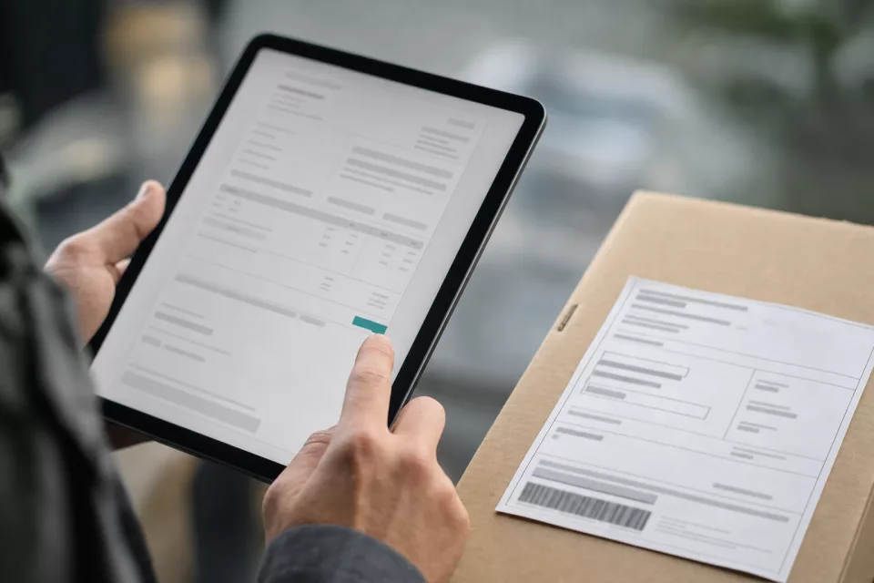 Gros plan sur des mains tenant une tablette montrant une facture électronique structurée à côté d’une étiquette papier sur un colis, illustrant la coordination des données et des systèmes autour de la facture électronique.
