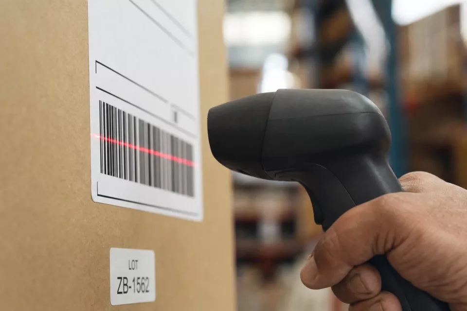 Gros plan macro d’un lecteur code-barres industriel scannant l’étiquette d’un colis en carton dans un entrepôt, une image évocatrice de la gestion de stock et de la traçabilité.