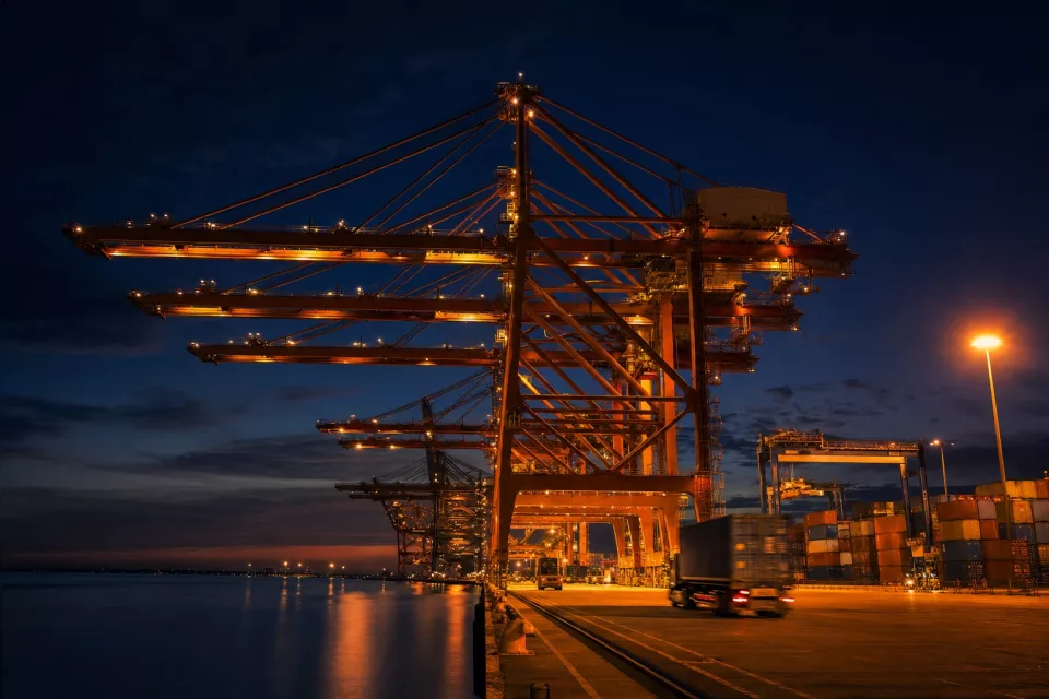 Photographie nocturne en pose longue de portiques de quai sur un terminal à conteneurs, évoquant l’optimisation transport et la coordination des flux logistiques.