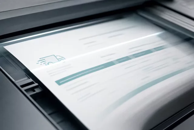 Gros plan éditorial d’une feuille papier entrant dans un scanner de production, avec une fine lumière de capture, illustrant la dématérialisation documentaire dans la supply chain.