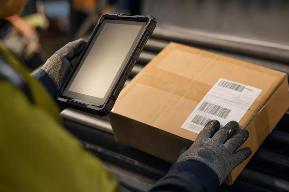 Gros plan sur les mains gantées d’un opérateur tenant une tablette durcie en scannant un colis sur une ligne de tri, dans un contexte de promesse de disponibilité ATP omnicanale.